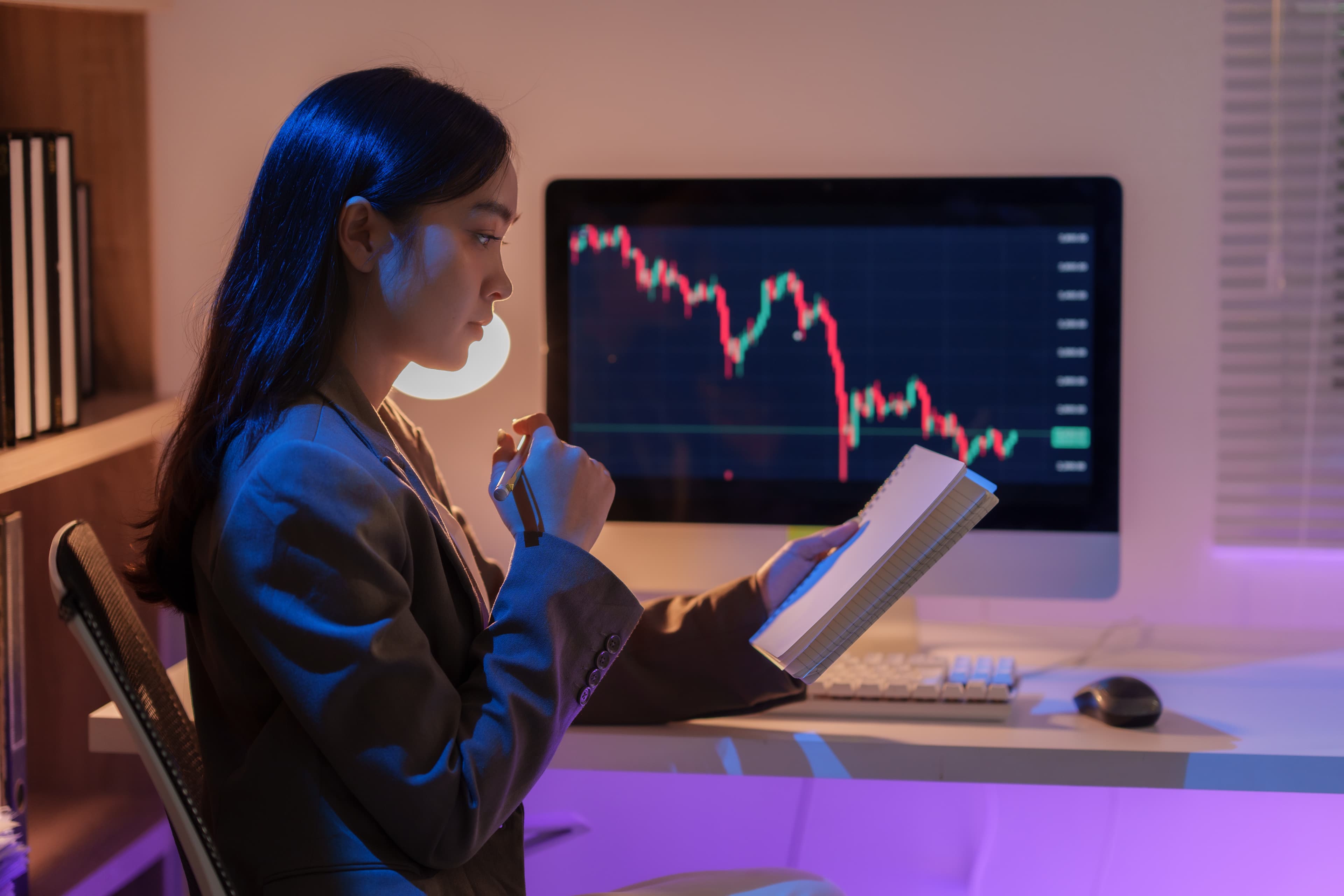 Trader escribiendo en su journal de trading frente al monitor con gráficas de futuros
